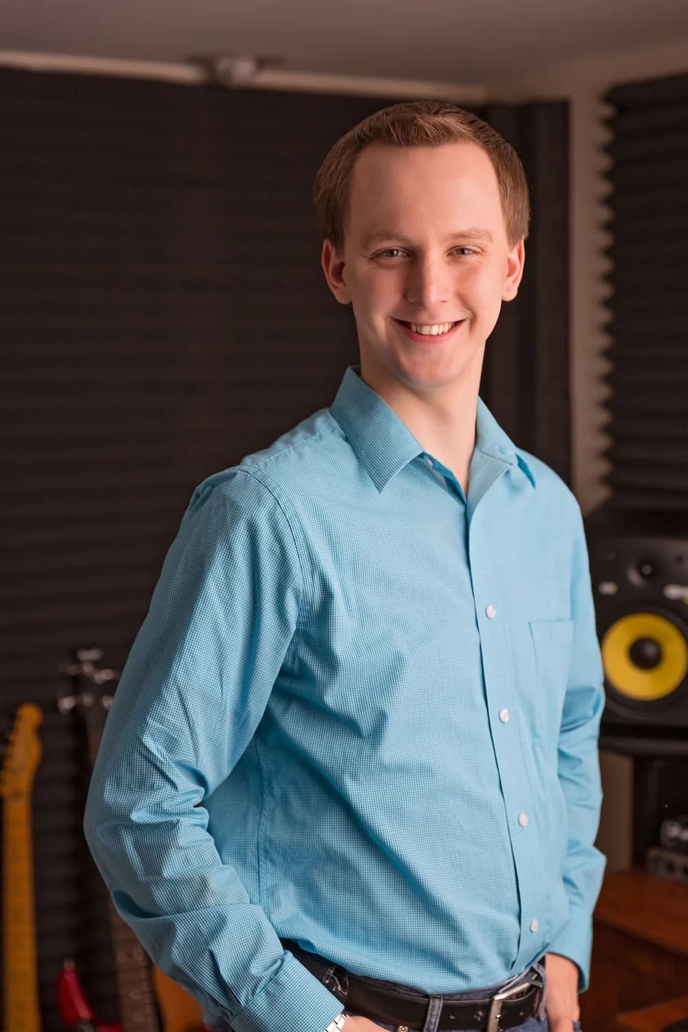 Stephen Forester smiling in a blue shirt in the studio, with guitars and a KRK monitor behind him